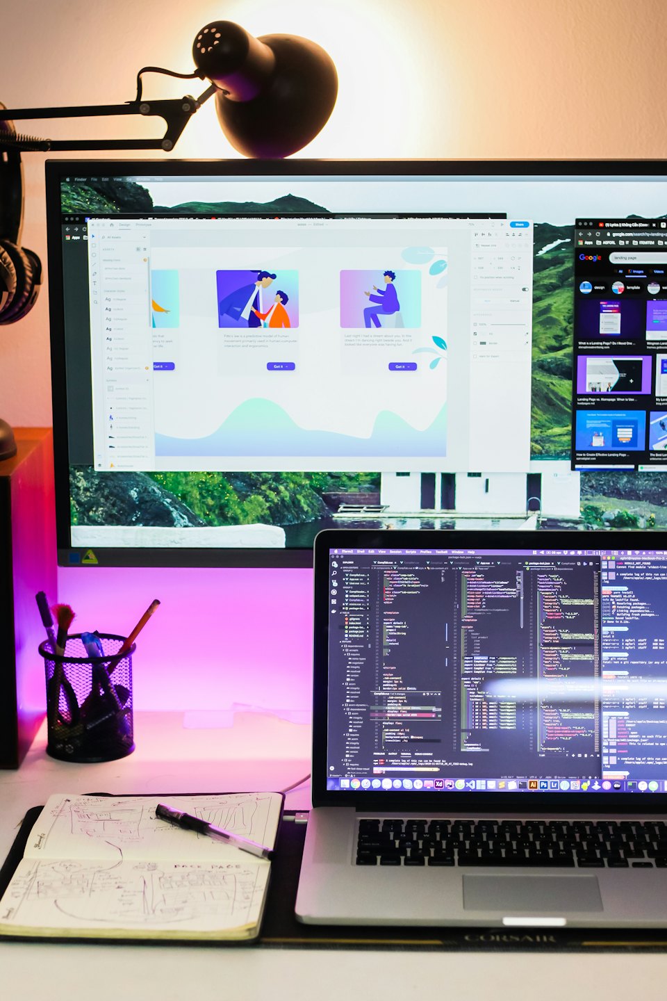 Meja modern dengan monitor komputer, laptop, dan buku catatan.